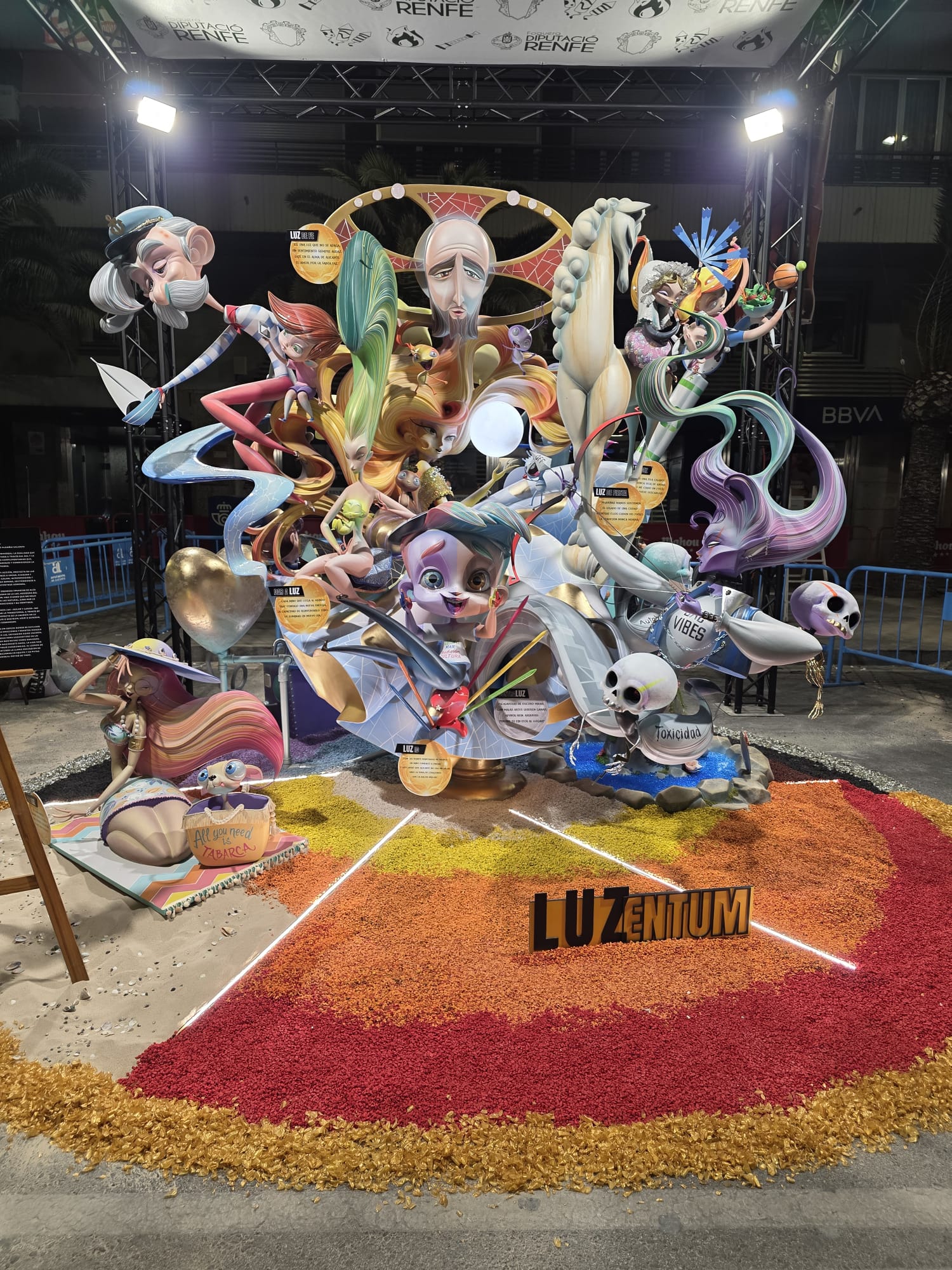 Vista frontal de la Hoguera Infantil Diputació-Renfe 2026, donde se aprecia la composición completa del monumento y el trabajo realizado en la decoración del suelo. La escena combina piedras decorativas en una amplia gama cromática —rojos, naranjas, amarillos, marrones y negros— creando diferentes zonas de color que delimitan cada ambiente. La iluminación LED en tonos fríos y cálidos aporta profundidad y dirige la mirada hacia el conjunto escultórico, mientras que el hidrogel azul y transparente y los detalles paisajísticos integran todos los elementos en una composición dinámica, moderna y llena de contrastes. El resultado es un monumento en el que materiales, iluminación y color trabajan conjuntamente para reforzar el mensaje artístico de la hoguera.
